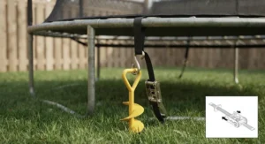 A close-up photograph of a black polyester ratchet strap from the anchor kit, looped over the top rail of the galvanized trampoline frame, threaded through the ratchet mechanism, and connected with a J-hook to the installed yellow spiral anchor stake in the grass, showing high tension.