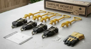 A flat lay photograph on a textured grey surface showing all the contents of the trampoline anchor kit: four bright yellow spiral steel anchor stakes, four black heavy-duty ratchet straps with J-hooks, a pair of yellow and grey work gloves, a small bag of hardware, and an instruction manual, with the branded packaging box in the background.