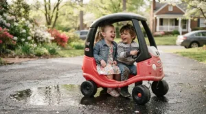 Siblings laughing as they drive a bright yellow two-seater electric ride-on car for two kids past spring puddles.